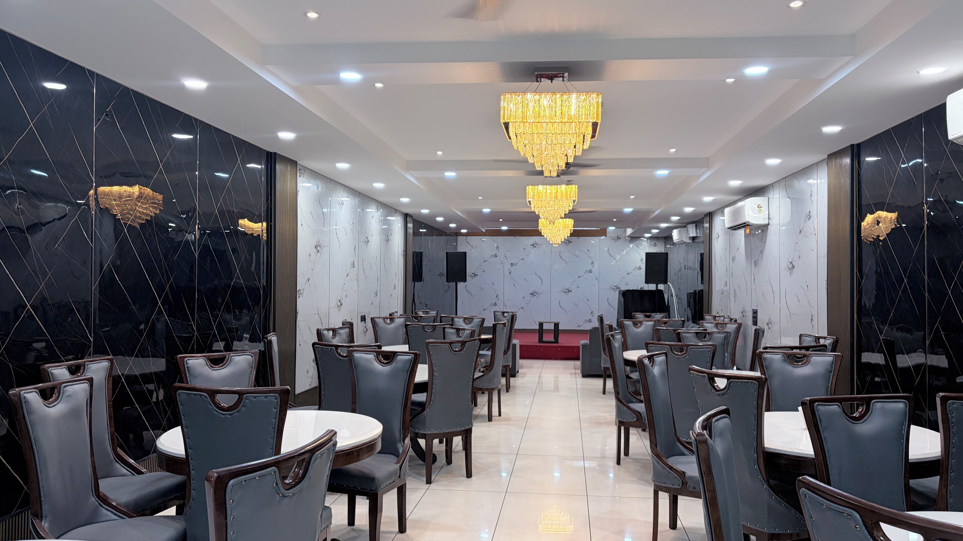 A spacious dining area with multiple tables and chairs, a large chandelier, and marble walls.