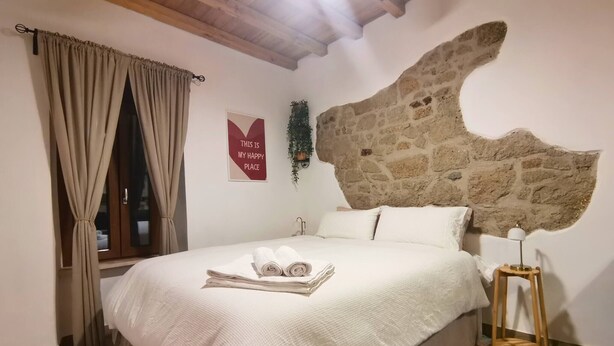 Ein Schlafzimmer mit Stehwänden, einem Bett mit weißer Bettwäsche, Holzboden, einem Fenster mit Vorhängen und einer Wandpflanze.