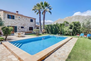 Ein Swimmingpool mit einer Steindeck, ein Essbereich mit Gartenmöbeln und ein Steinhaus mit blauen Fensterläden.