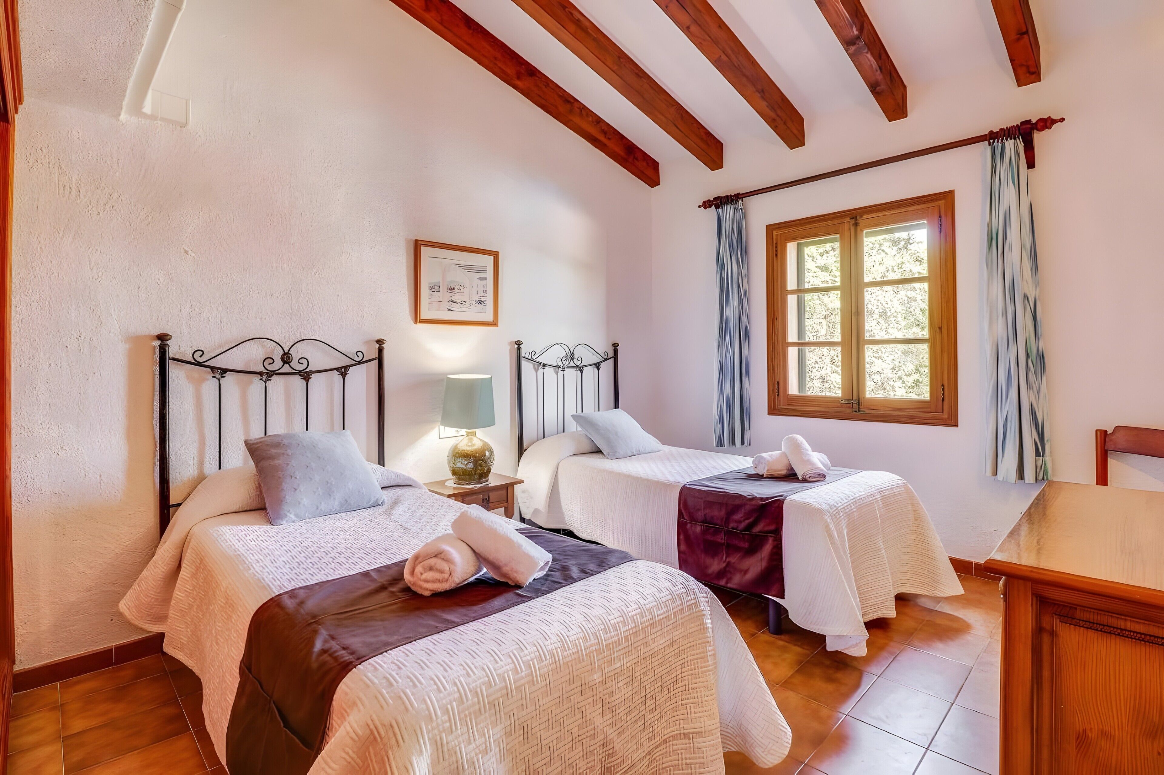 Una habitación con dos camas, una ventana con cortinas, una lámpara y un cuadro en la pared.