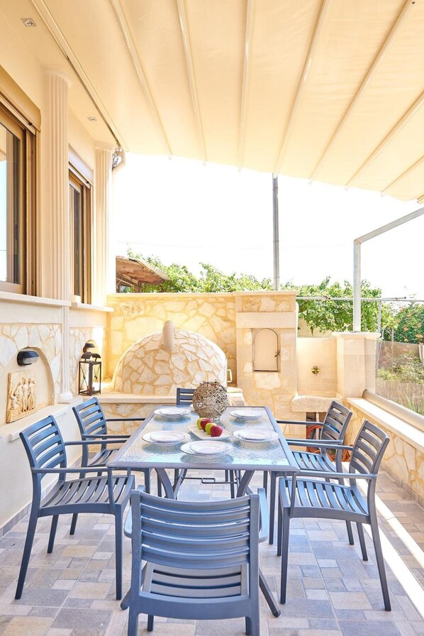 An outdoor dining area with a stone fireplace, a glass table, and blue chairs. - Villa 'Vlite' with Sea View, Wi-Fi and Air Conditioning (Souda)