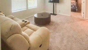 Uma sala de estar com um sofá modular bege, uma mesa de centro redonda e uma televisão de tela plana na parede.