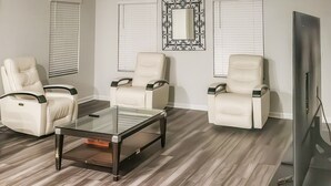 Uma sala de estar com uma mesa de centro de vidro, poltronas de couro branco, uma televisão de tela plana e um espelho decorativo na parede.