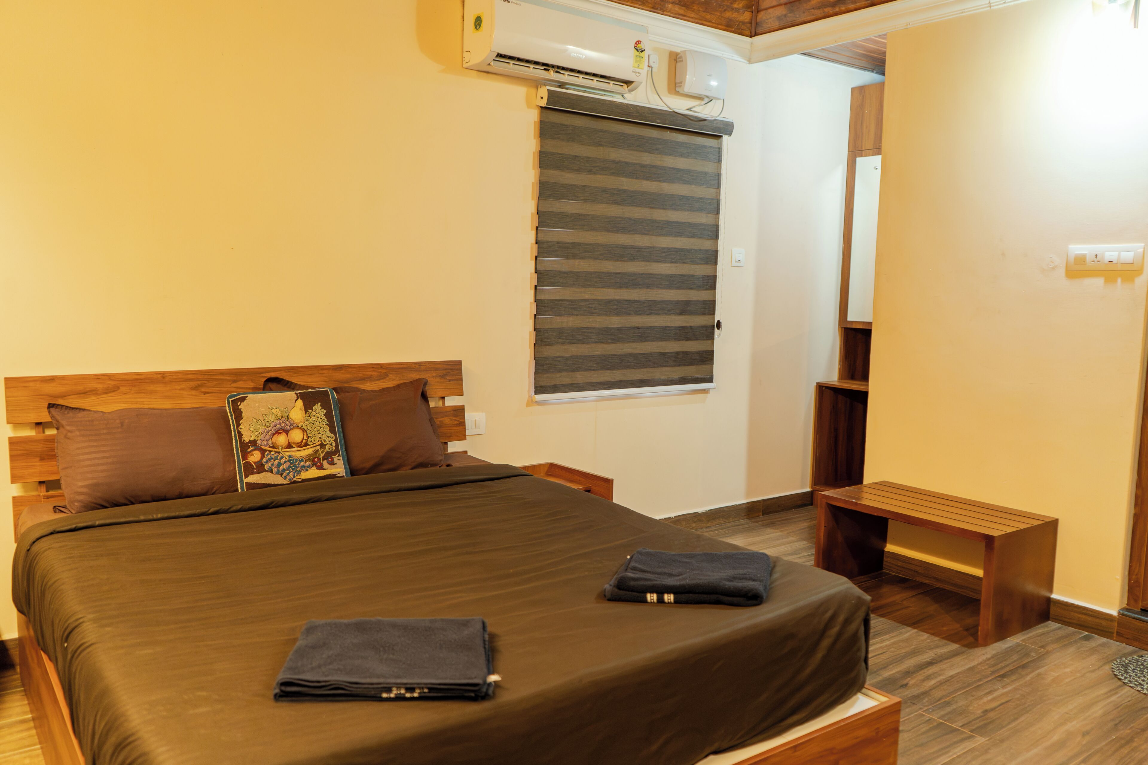 A bedroom with a bed, pillows, a bedside table, a bench, an air conditioning unit, and a window with blinds.