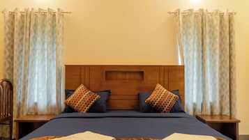 Habitación de hotel con cabecera de madera, dos almohadas, cama con cubrecama azul, dos mesitas de noche y cortinas.