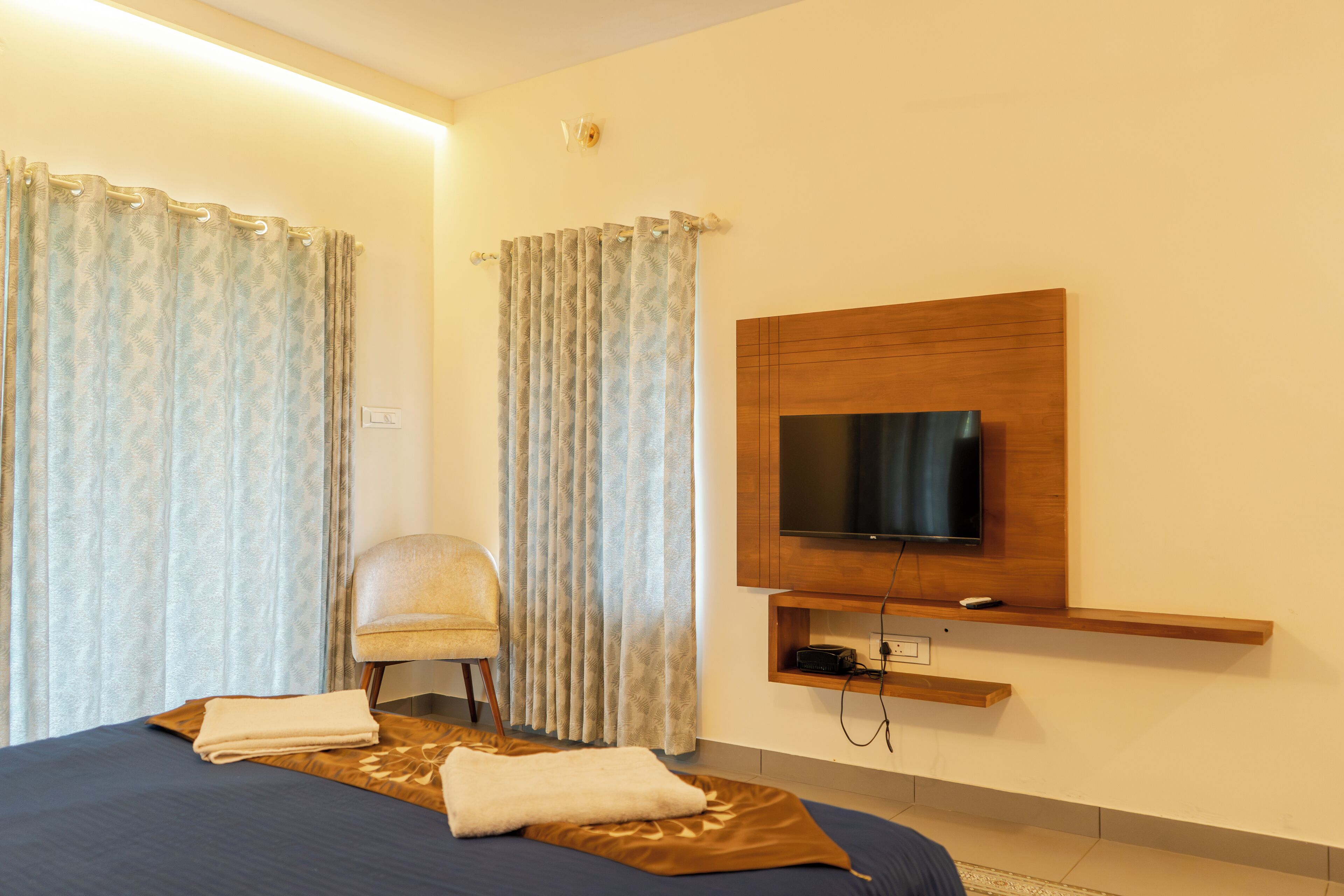 A hotel room with a bed, a chair, a television mounted on a wooden panel, and curtains.