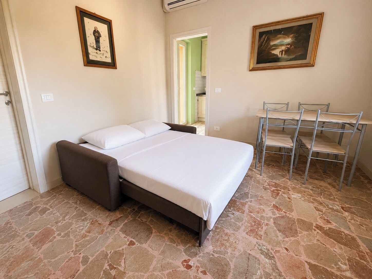 A bedroom with a bed, a desk with chairs, and a door leading to another room.