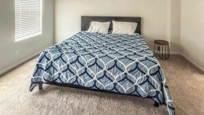 Un dormitorio con cama, ventana con persianas, un taburete pequeño y suelo alfombrado.