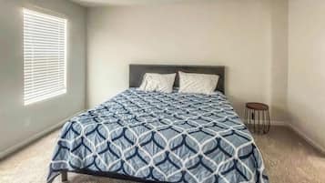 A bedroom with a bed, a window with blinds, a small stool, and a carpeted floor.