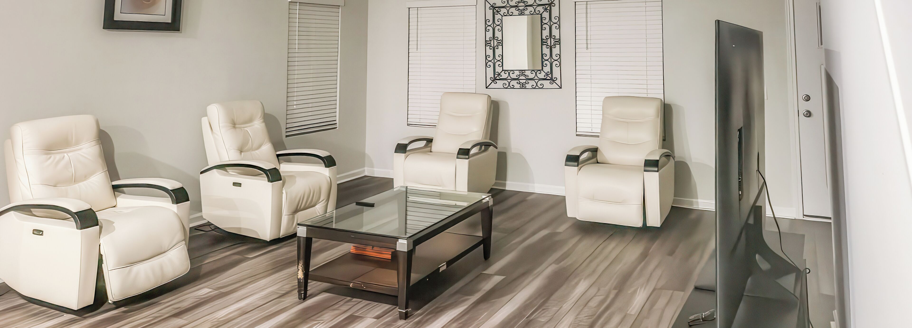 A room with a glass coffee table, four white leather chairs, a flat-screen TV, and wooden flooring.