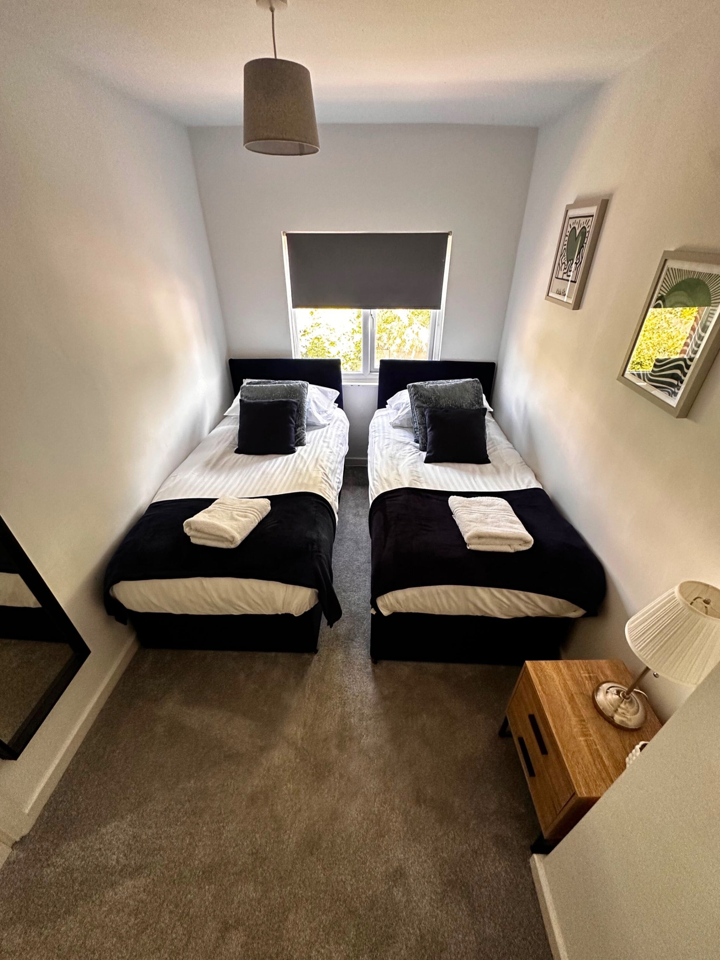 Une chambre avec deux lits, une fenêtre, une lampe et une petite table de chevet en bois.