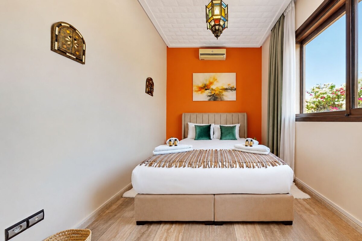 Habitación de hotel con cama, un cuadro, aire acondicionado y ventana con vistas a flores.