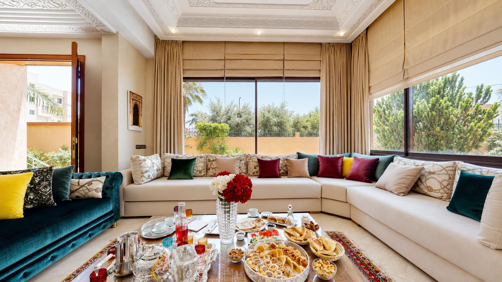 A spacious living area with a sofa set, a dining table with food and drinks, and large windows offering a view of greenery.