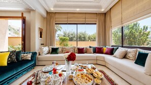 Amplia sala de estar con un conjunto de sofás, una mesa de comedor con comida y bebidas, y grandes ventanales con vista a follaje.