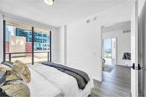 A modern hotel room with a large bed, a view of a cityscape through floor-to-ceiling windows, and a separate entrance leading to a balcony. - Luxury 2BR Retreat - Water Views, Pool & Walk to Bayside (Miami)