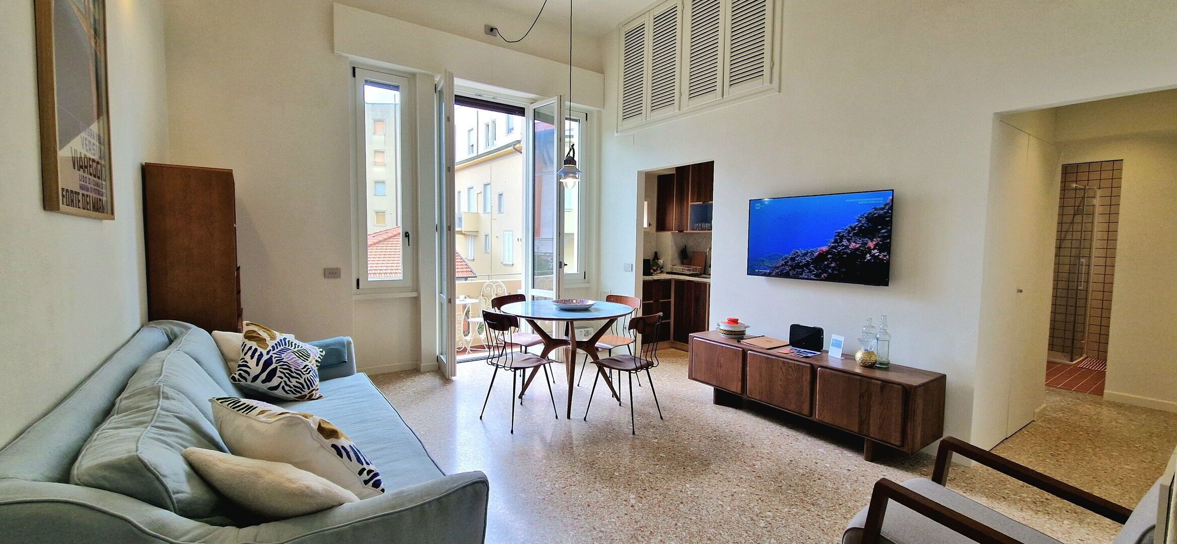 A living room with a sofa, a dining table with chairs, a TV mounted on the wall, and a kitchen area.