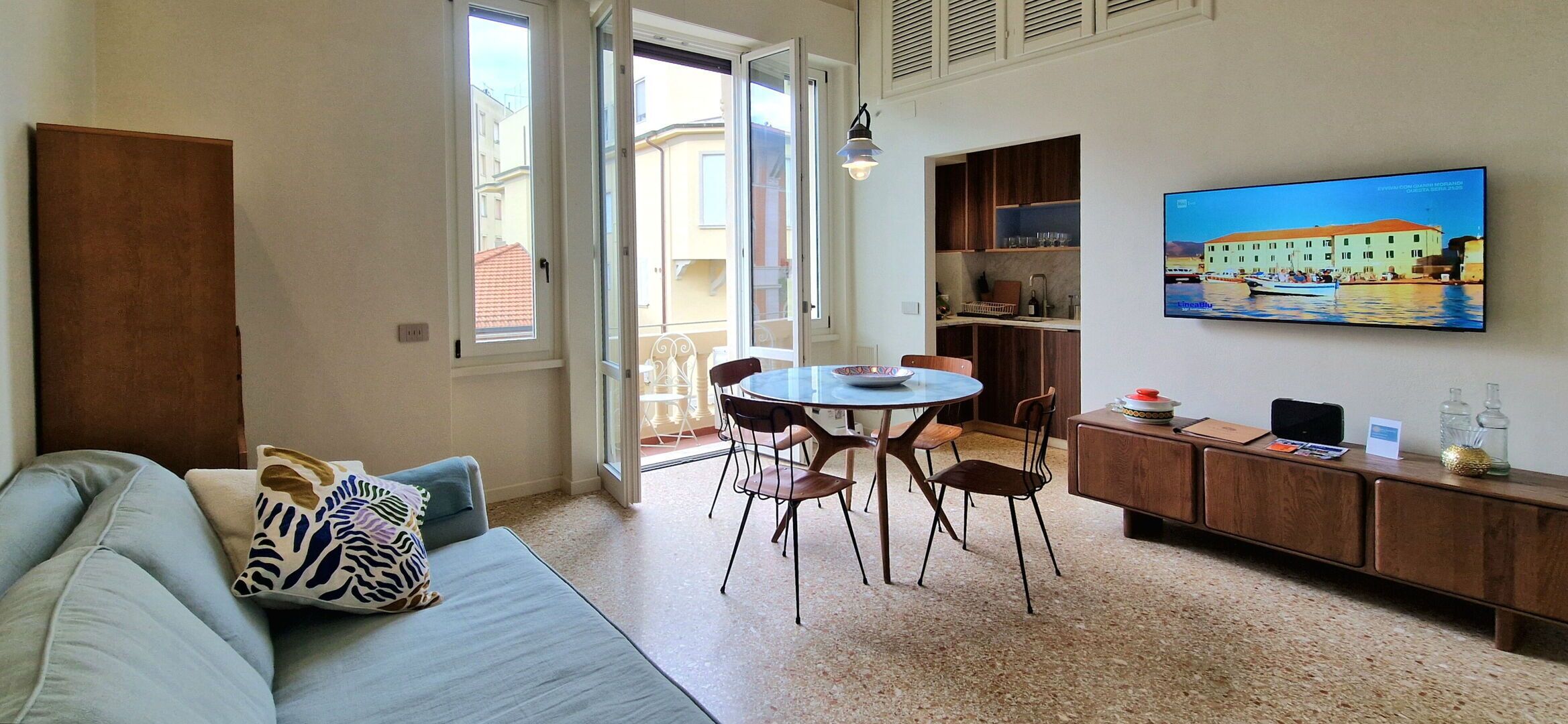 A living room with a sofa, a dining table with chairs, a kitchen area with a TV, and a balcony with a view.