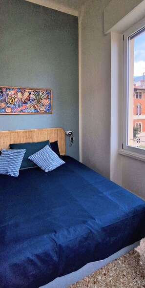 Ein Bett mit dunkelblauer Bettdecke und zwei Kissen, ein Gemälde an der Wand und ein Fenster mit Blick auf Gebäude.