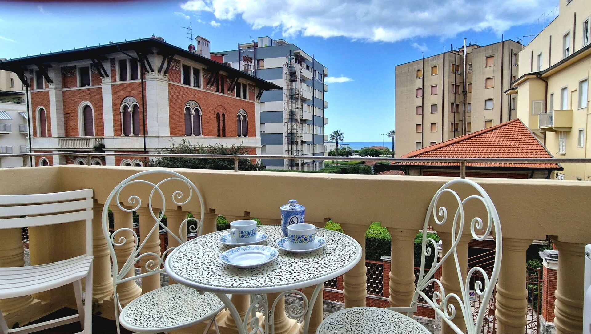 Un balcón con sillas de metal blanco y una mesa dispuesta con platos azules y blancos, además de una tetera.