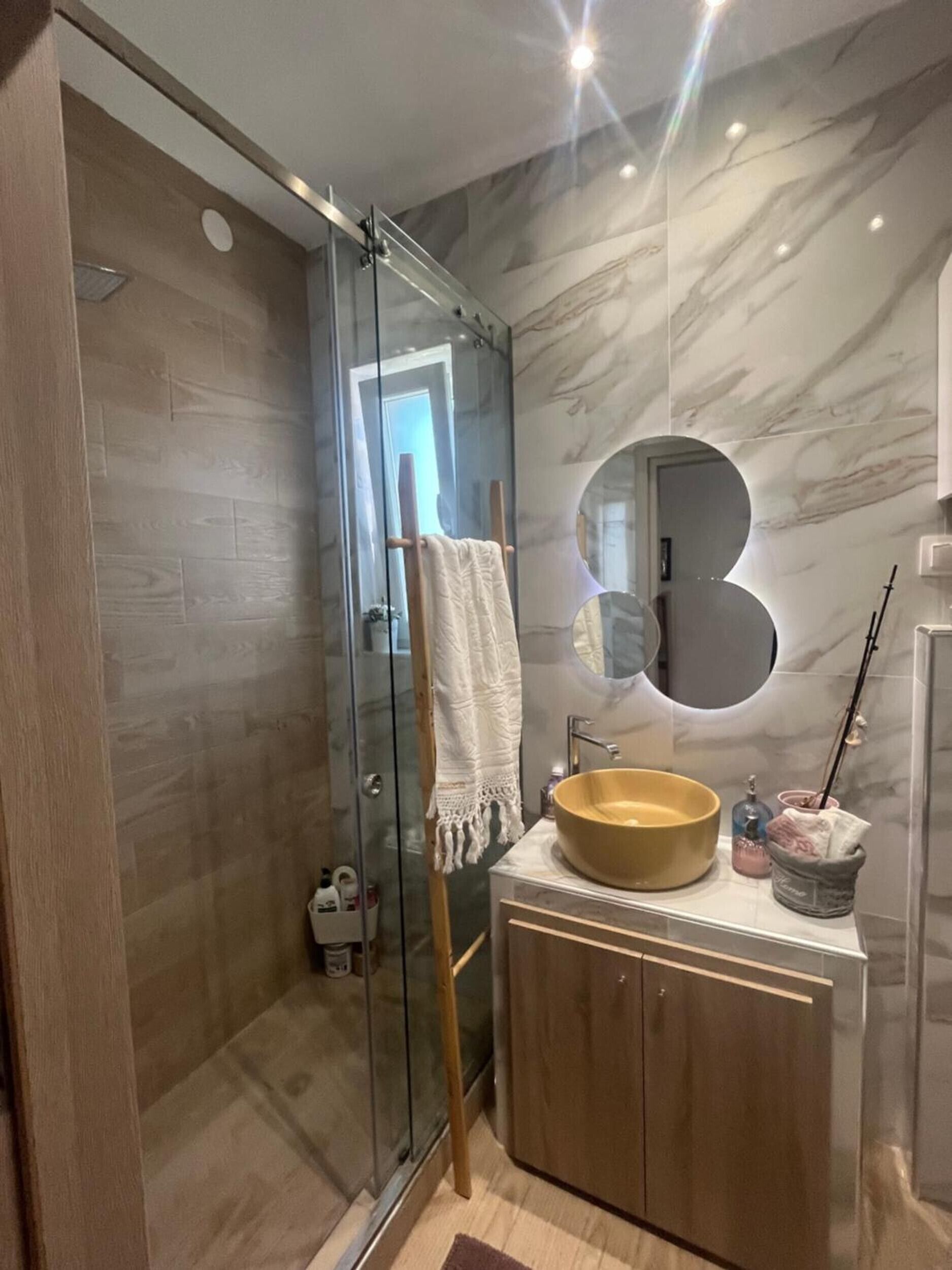 Une salle de bain moderne dotée d’une douche à parois de verre, d’un meuble-lavabo en bois avec un vasque rond et d’un miroir à cadre circulaire.