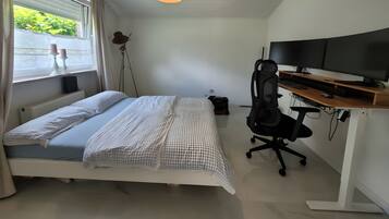 Un dormitorio con una cama, un escritorio con computadora, una silla y un ventanal con cortinas.