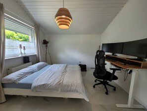 A bedroom with a bed, a desk with a computer, a chair, and a window with curtains.