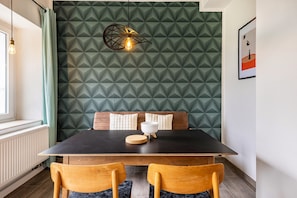 Ein Essbereich mit dunklem Tisch, zwei Holzsesseln, einer Wand mit Muster und einer Pendelleuchte. An der Wand hängt ein gerahmtes Bild.