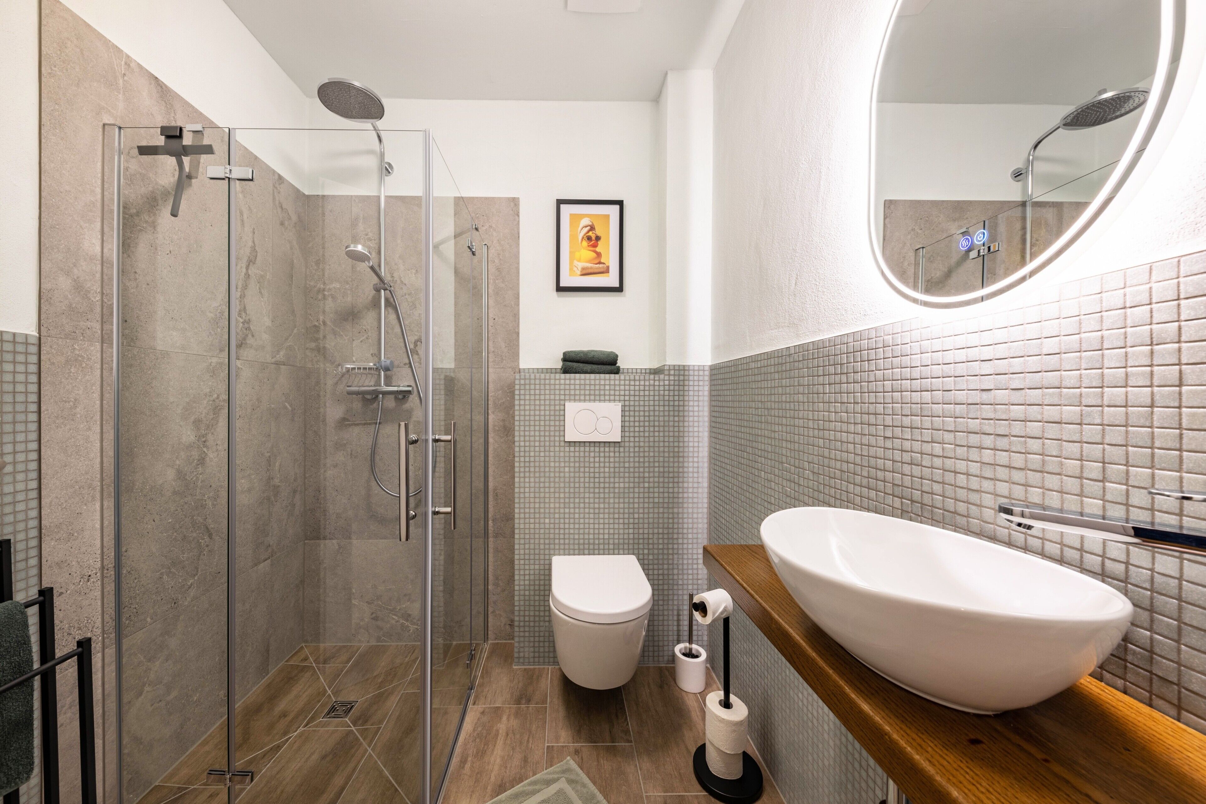 Et moderne badeværelse med en brusekabine i glas, et hvidt toilet og en stor, oval håndvask med træbordplade.