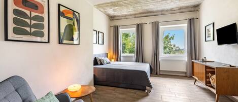A hotel room with a bed, a sofa, a small table, a TV, and artwork on the walls.