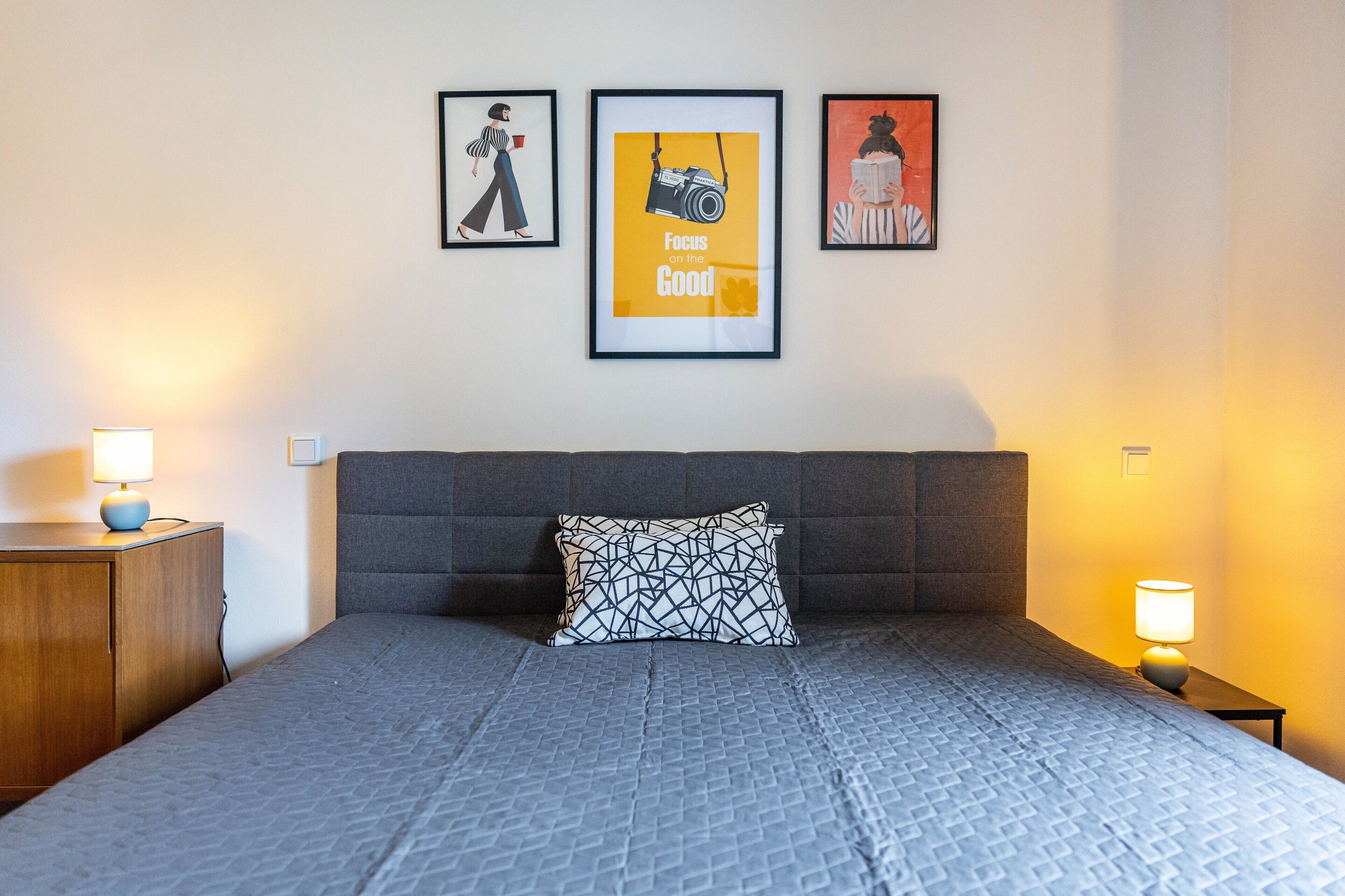 Un lit bien fait, avec une tête de lit grise et une couvre-lit à motifs, flanqué de deux tables de chevet surmontées de lampes, et trois œuvres d’art encadrées accrochées au mur.