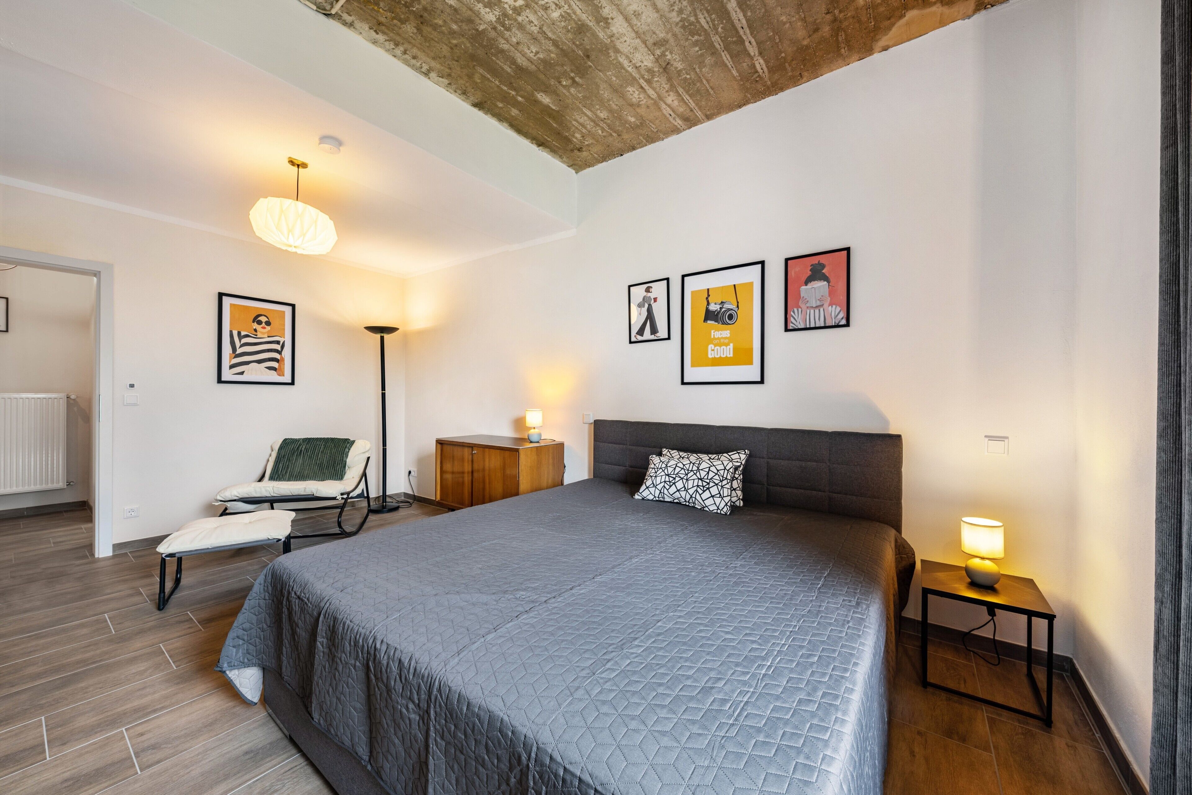A bedroom with a large bed, a chair, a nightstand, and a floor lamp. There are framed pictures on the wall.