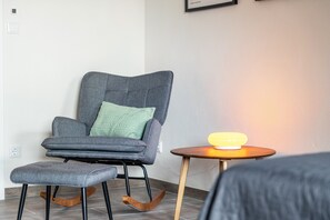 Ein modernes Zimmer mit einem grau gepolsterten Sessel, passendem Hocker und einem kleinen Holfnachtkästchen mit einer gelben Lampe.