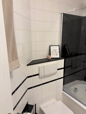A bathroom with white tiles, a black bathtub, a glass shower enclosure, and a framed botanical print on the wall. - Charming apartment surrounded by greenery with great access to public transportation (Paderborn)