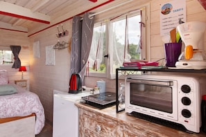 A kitchenette with a microwave, a kettle, and a small oven. There is a window with curtains and a view of greenery outside.