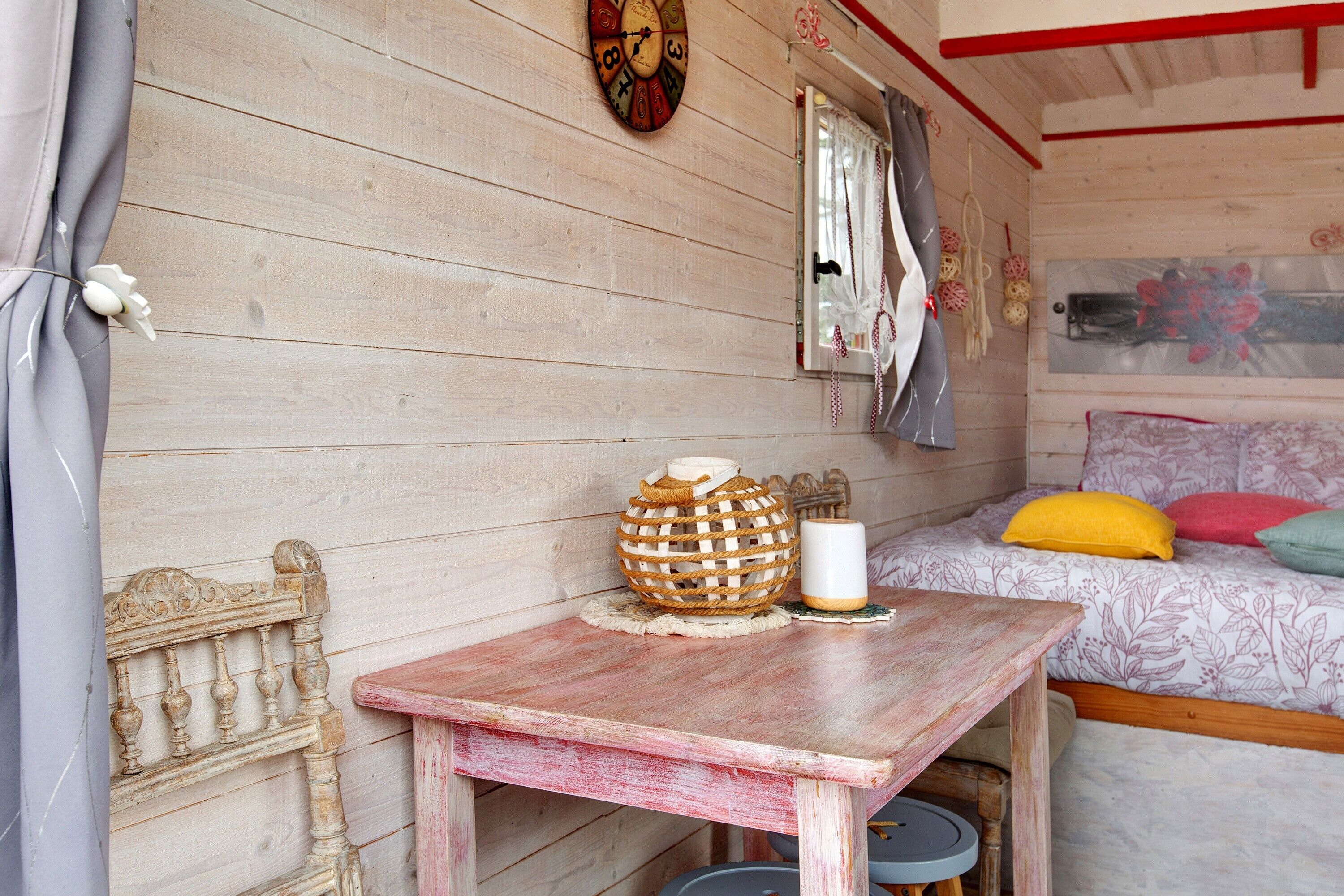 Ein kleines Zimmer aus Holz mit einem Bett, einem Tisch mit Kerze und einer dekorativen Wanduhr.