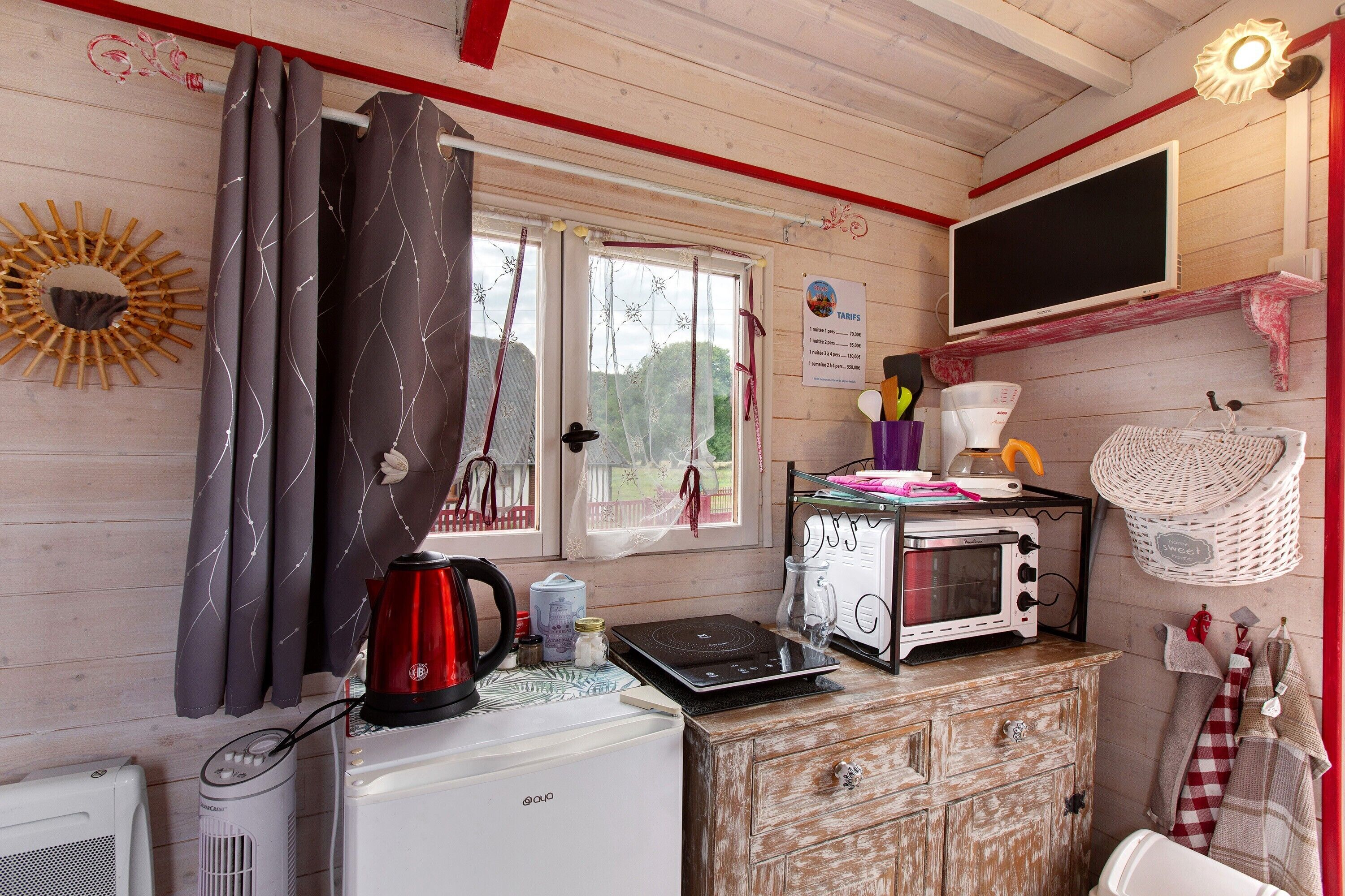 Une cuisine compacte équipée d’un micro-ondes, d’une bouilloire et d’un grille-pain, le tout posé sur un meuble en bois. On aperçoit une fenêtre avec des rideaux et un téléviseur fixé au mur.