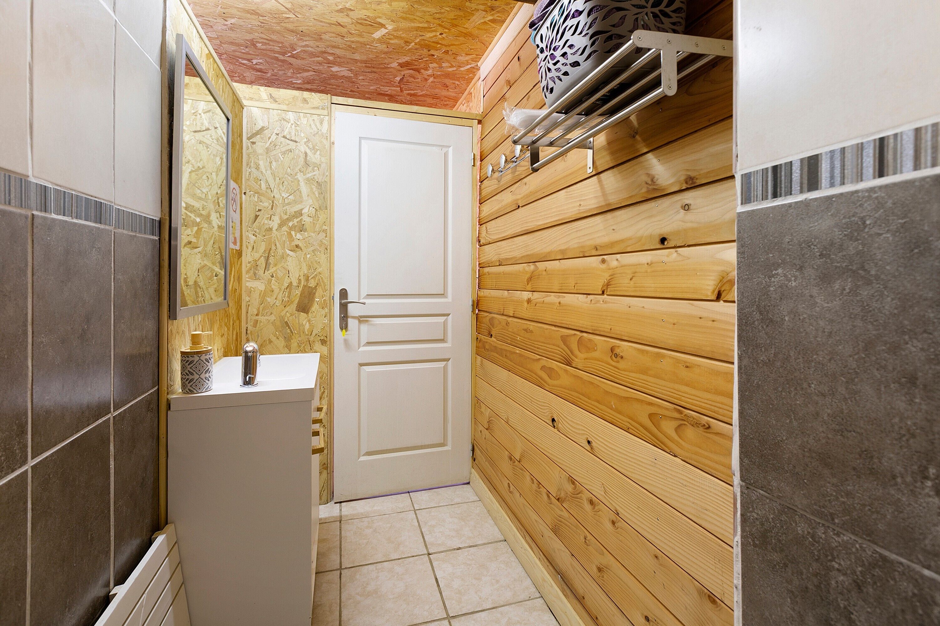 Ein schmales Badezimmer mit weißer Tür, Holzwänden, Fliesenboden und einem kleinen Waschbecken mit Seifenspender.