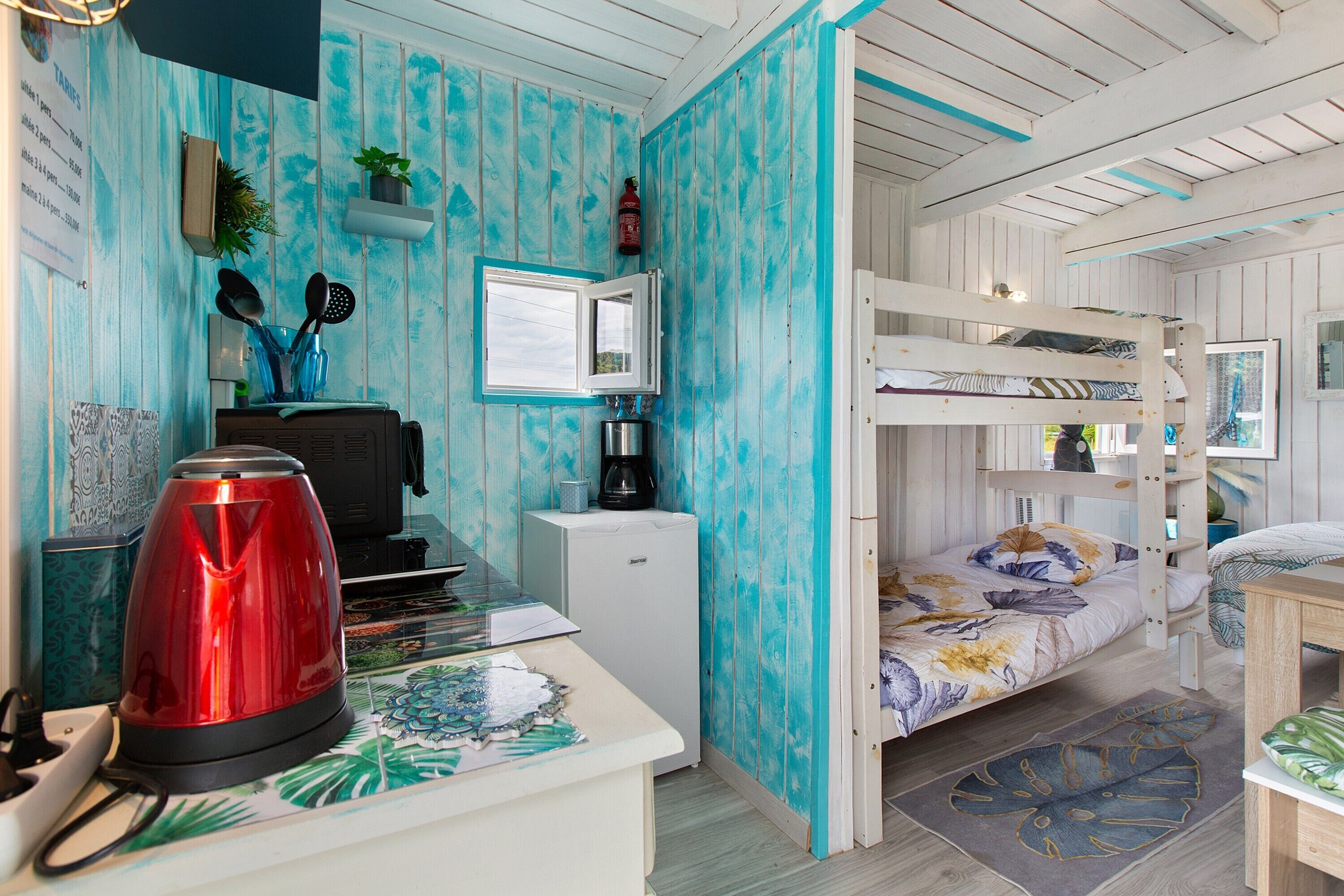 Une pièce compacte comprenant un lit superposé, une petite kitchenette et un bureau avec une bouilloire rouge.
