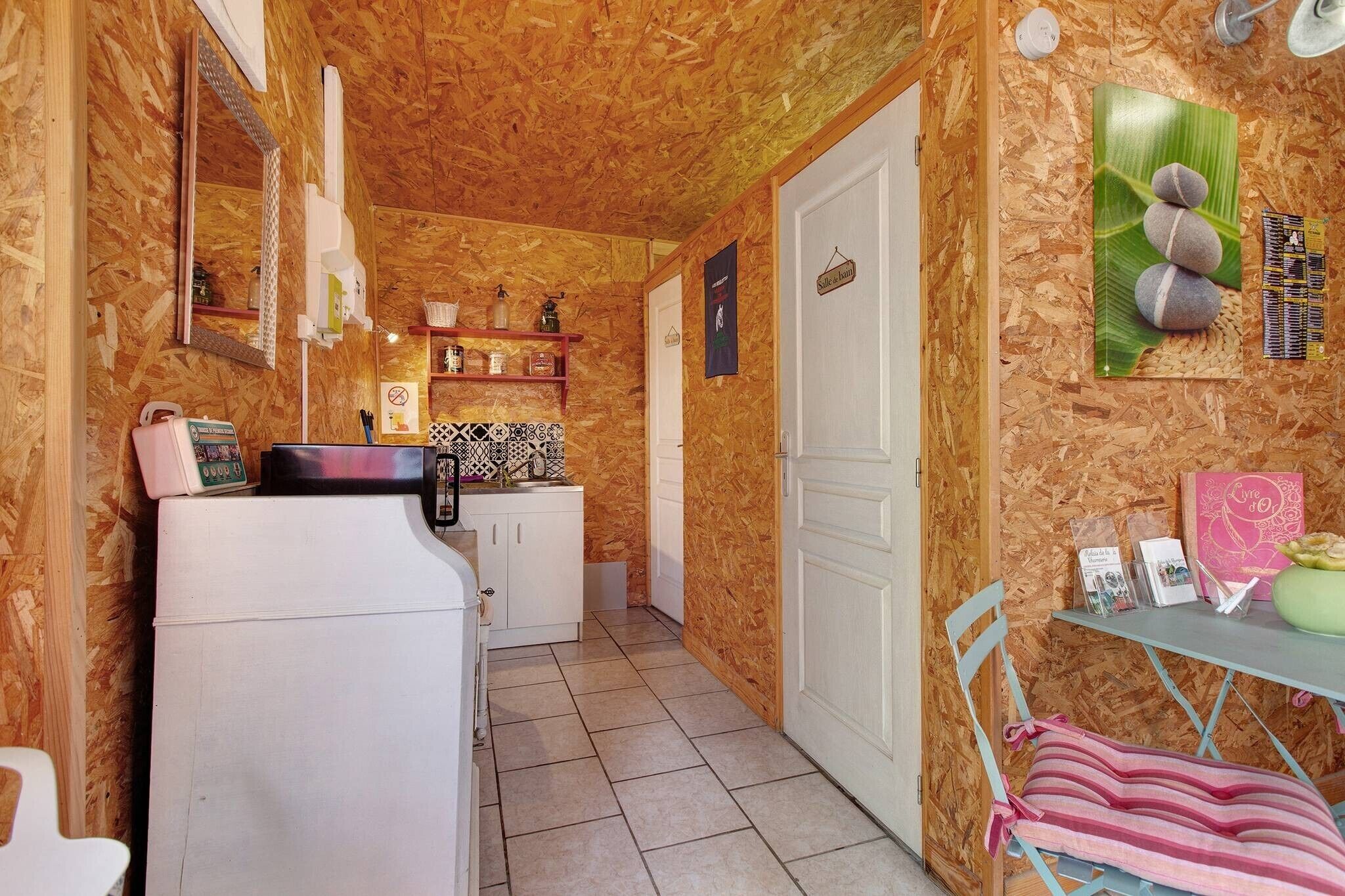 A compact kitchen with a refrigerator, sink, and a small countertop. There is a door leading to another room and a shelf with various items.
