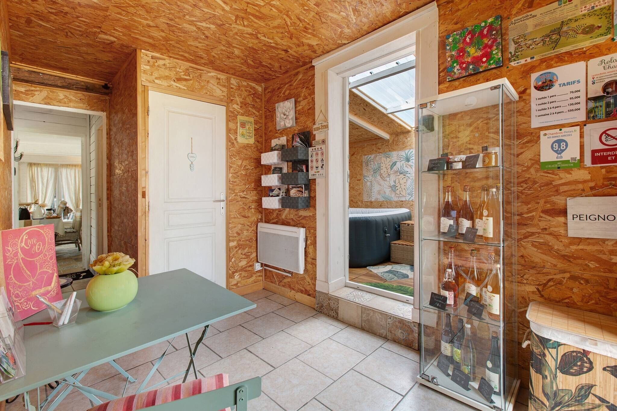 A room with a wooden ceiling, a white door, a table with a vase, a shelf with books, and a glass display case with bottles.