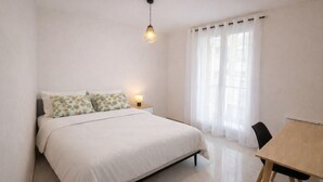 Ein Schlafzimmer mit Bett, Nachttisch mit Lampe, Fenster mit Vorhängen und kleinem Tisch mit Stuhl.