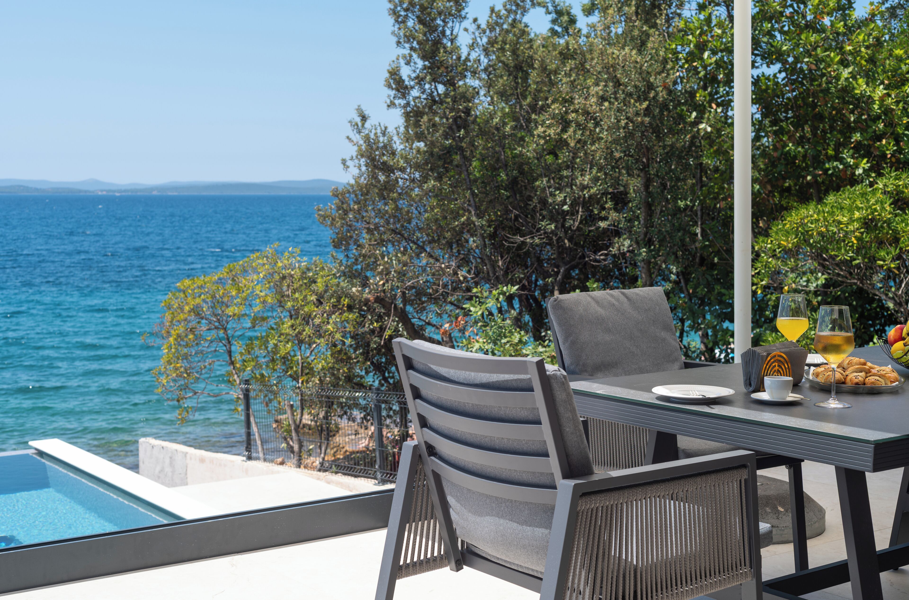 An outdoor dining area with a view of the sea, a table set with a plate, glasses, and a basket of bread.
