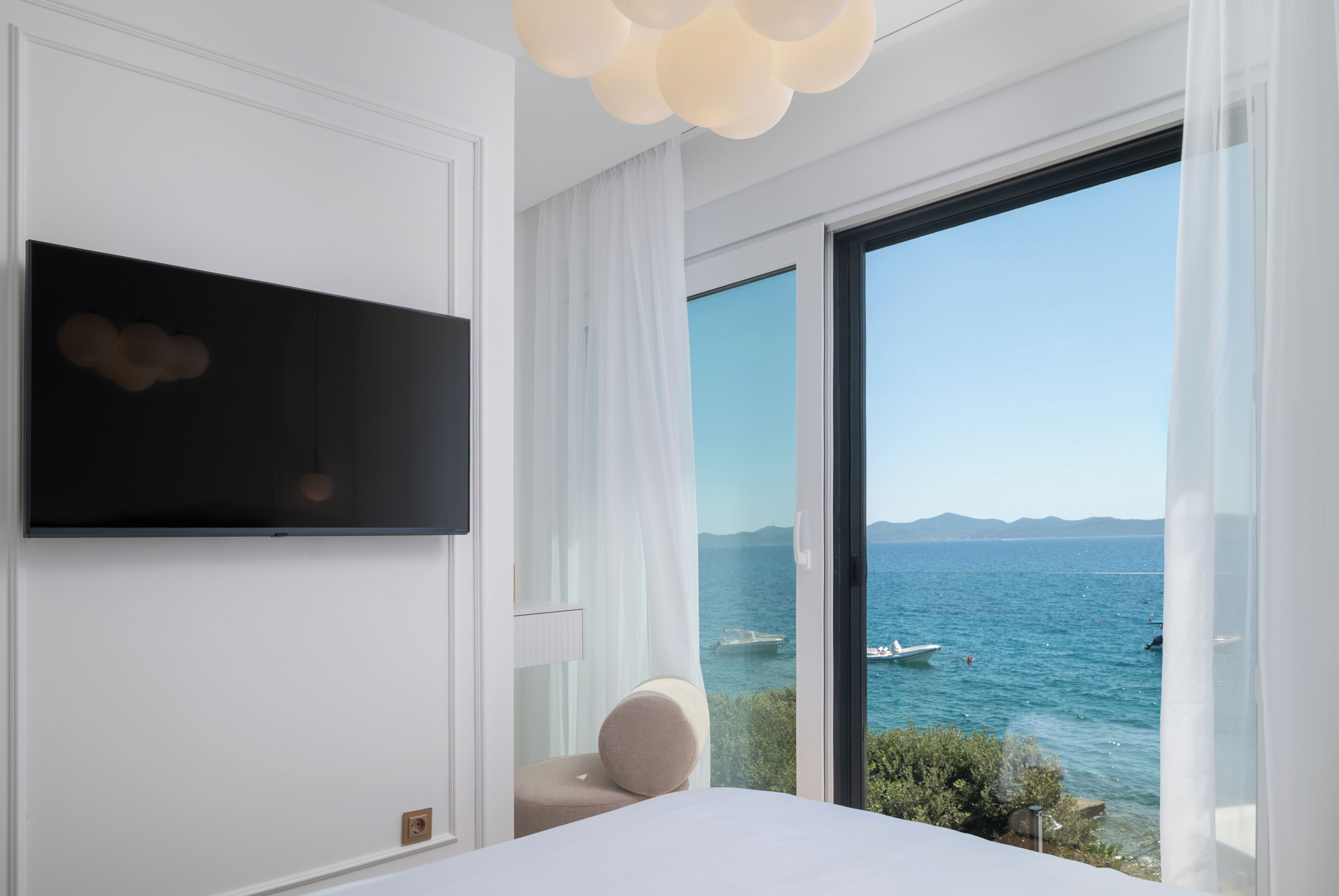 A modern hotel room with a large window, a flat-screen TV mounted on the wall, and a bed with a white headboard.