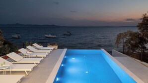A swimming pool with lounge chairs and tables, overlooking a body of water with boats. - Villa Silvio (Kožino)