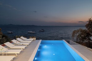 A swimming pool with lounge chairs and tables, overlooking a body of water with boats. - Villa Silvio (Kožino)