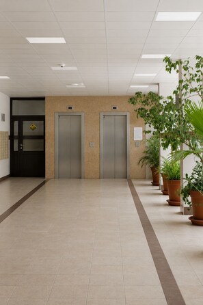 A hallway with two elevators, a door with a hazard sign, and potted plants. - Lumina 2-room Apartment near Metro  (Warszawa)