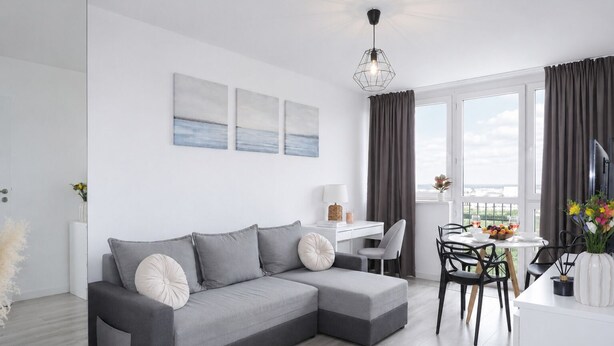 A modern living room with a grey sectional sofa, a dining area with a table and chairs, and a view of the outdoors through large windows. - Lumina 2-room Apartment near Metro  (Warszawa)
