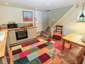 Una cocina compacta con microondas, horno y una encimera de madera. Una alfombra de patchwork colorida cubre el suelo, y una silla está colocada cerca de una mesa.