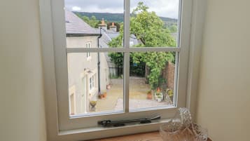 Une fenêtre donnant sur un jardin et des bâtiments, une table en bois avec un saladier en verre, et un rebord de fenêtre orné d’un petit objet décoratif.