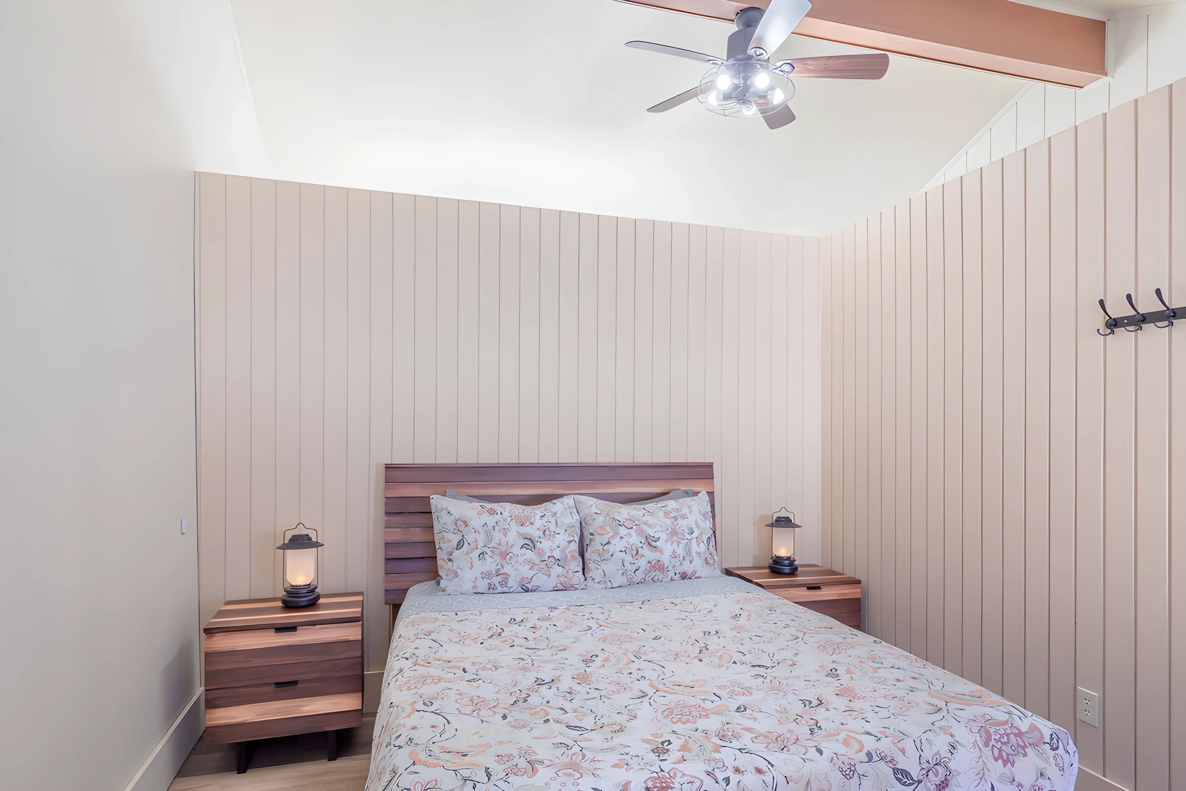 A bedroom with a bed, wooden headboard, two bedside tables with lamps, a ceiling fan, and a wall-mounted coat rack.